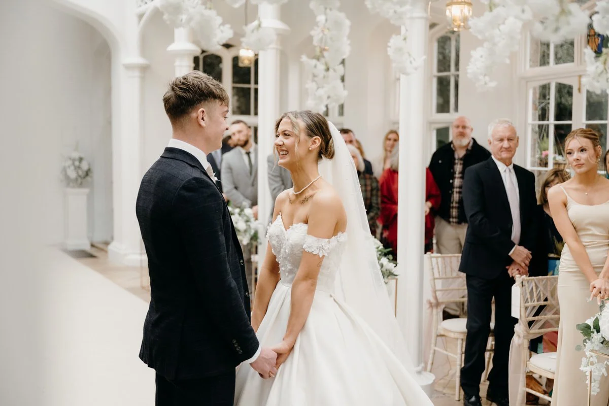 Bride and groom holding hands during their wedding ceremony in a bright, elegant venue with white flowers and guests watching photographed by Dorset wedding photographers pip&clo at St Audries Park, Taunton Somerset.