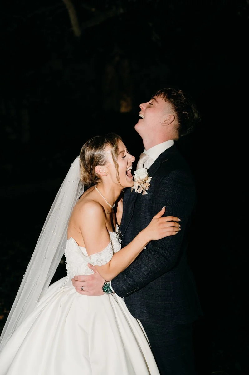 A bride and groom are smiling and laughing while holding each other during their wedding celebration, wearing traditional wedding attire, with the bride in a white gown and veil, and the groom in a dark suit.