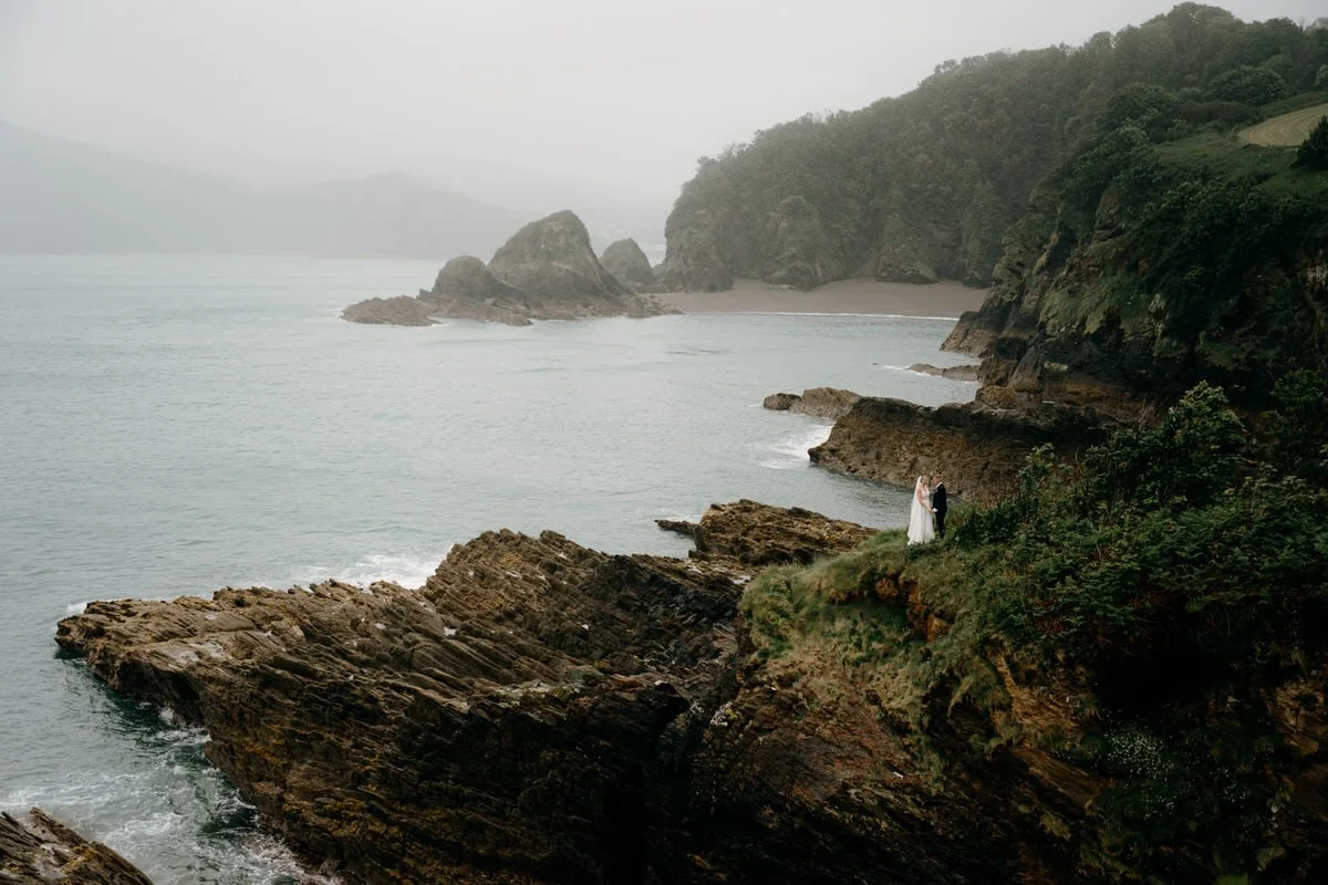 A bride and groom standing on a rugged cliffside overlooking the ocean on a cloudy day.