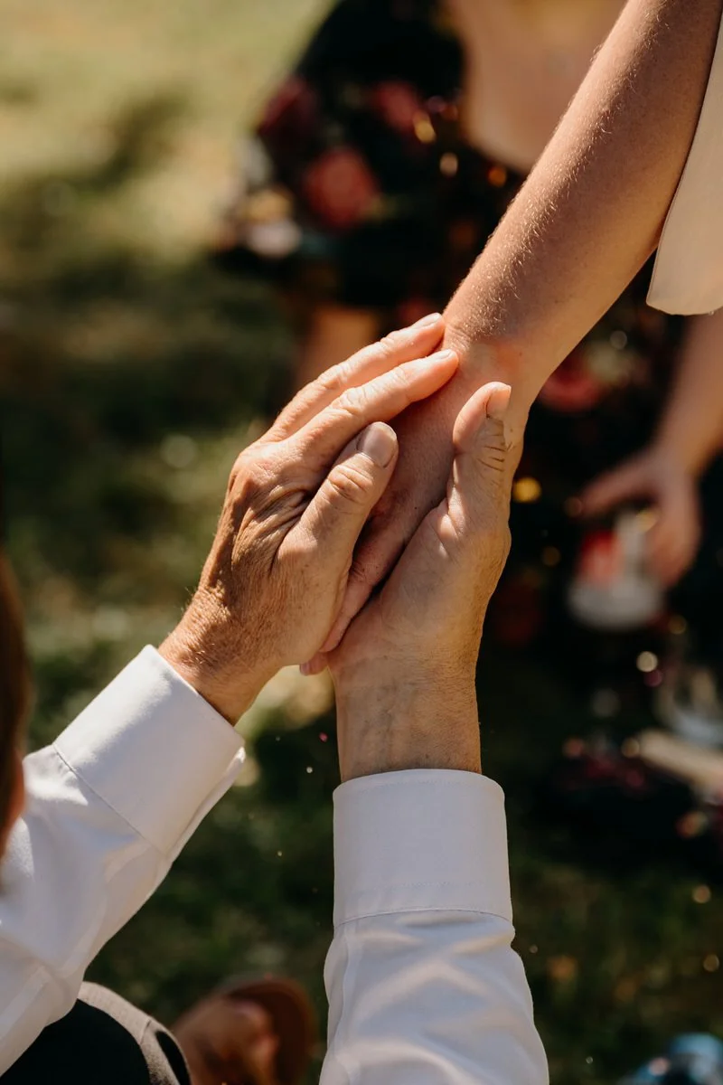 An elderly person gently holding a bride's hand during an outdoor wedding, with a blurred background of other people captured by Dorset Wedding Photographers pip&clo