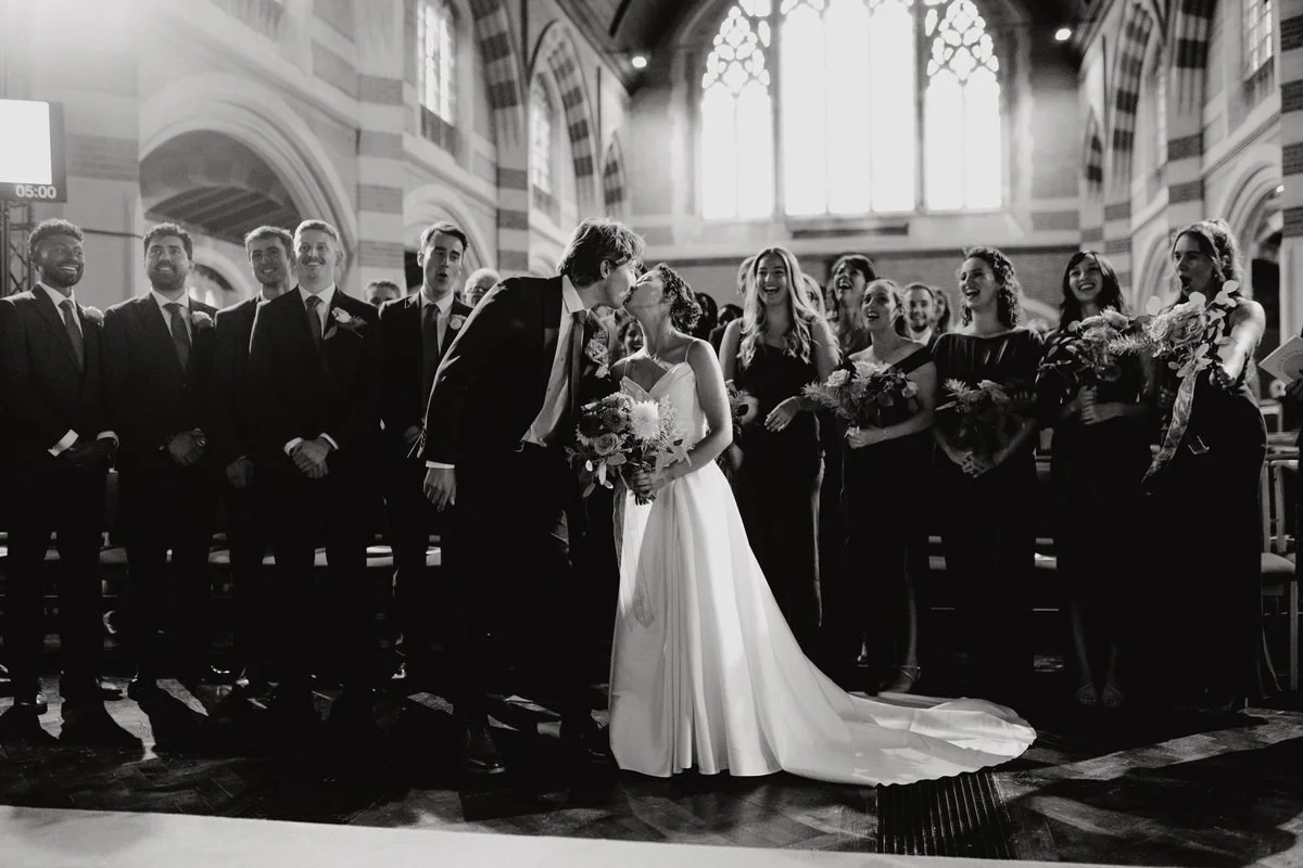 A black and white photo of a wedding ceremony inside a church, with a bride and groom kissing in the center, surrounded by bridesmaids and groomsmen.