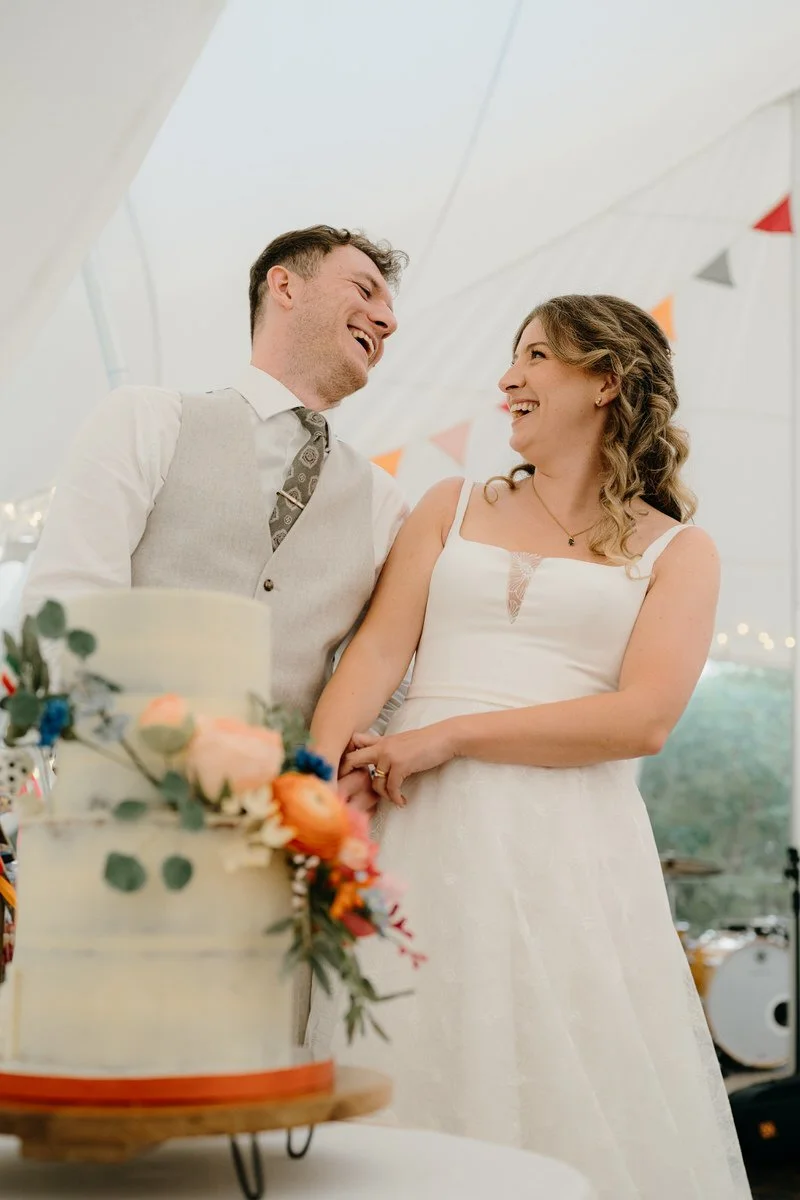 A bride and groom smiling and holding hands in a wedding reception tent with a decorated wedding cake in the foreground.