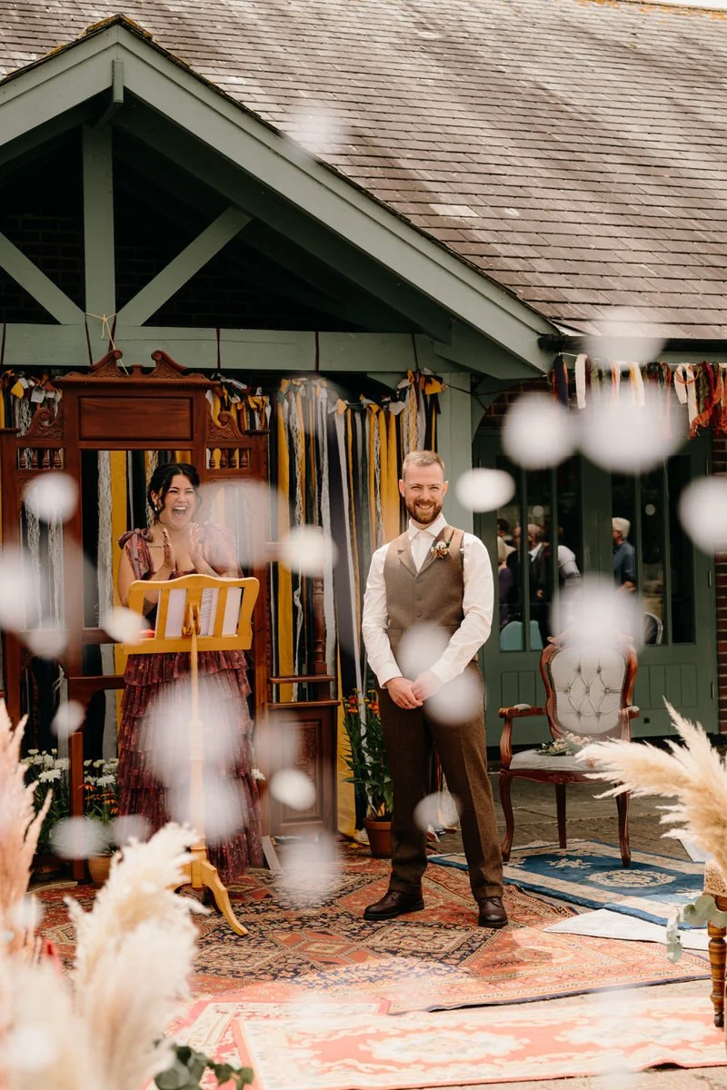A joyful wedding scene with a man in a brown vest and tie standing happily, and a woman in a red dress clapping with excitement, surrounded by vintage furniture and rustic decor outdoors.
