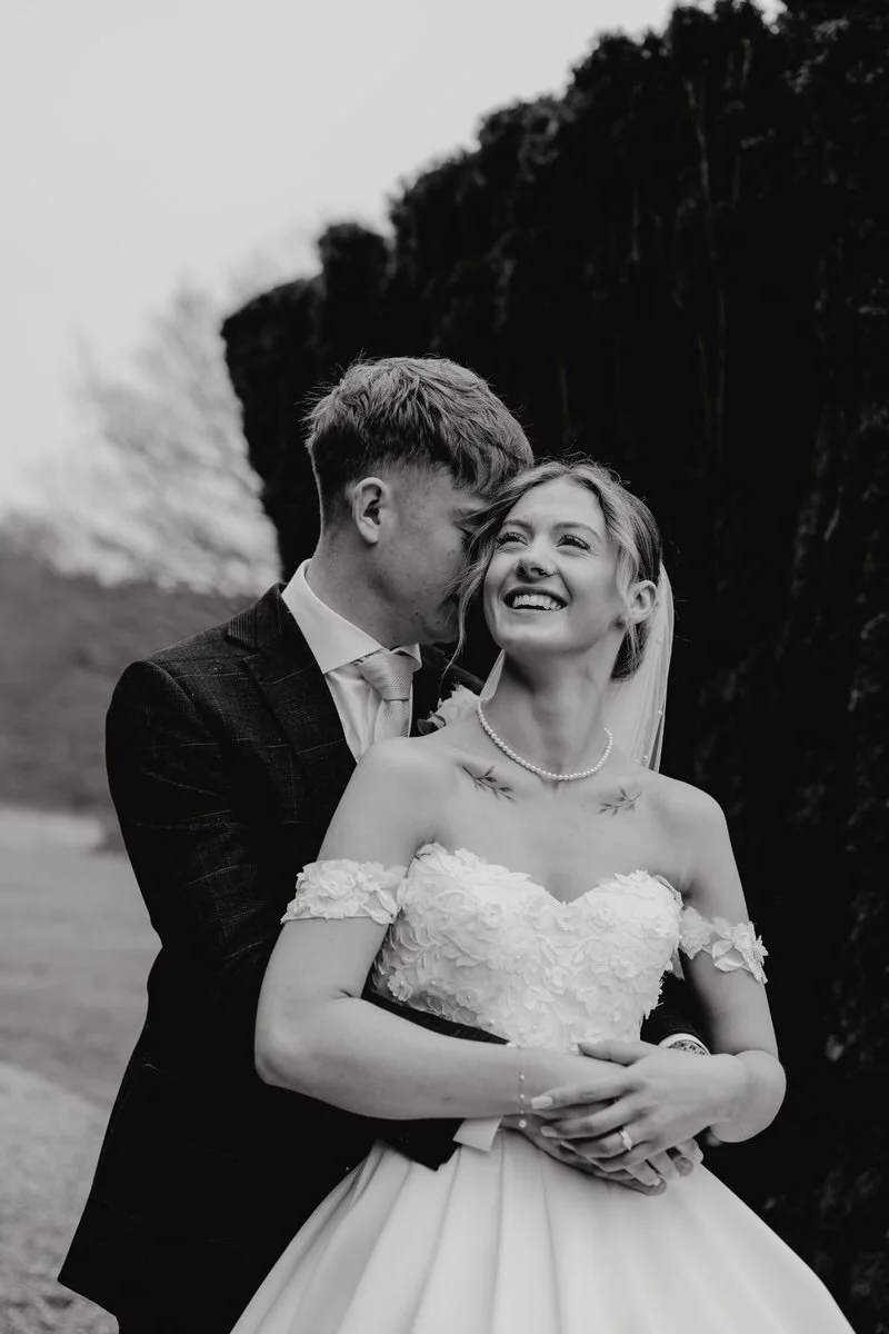 A black-and-white photo of a smiling bride and groom outdoors. The groom is kissing the bride on the cheek, and the bride is laughing with her eyes closed.
