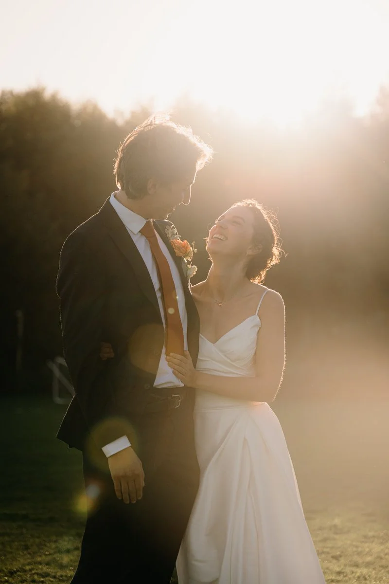A newlywed couple in wedding attire sharing a joyful moment outdoors at sunset, with the bride smiling up at the groom.