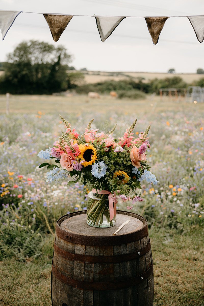 A vibrant bouquet of mixed flowers including sunflowers, roses, and other blooms in pastel colors arranged in a glass jar with a pink ribbon, placed on a wooden barrel in an outdoor setting with a field of wildflowers and a cloudy sky.