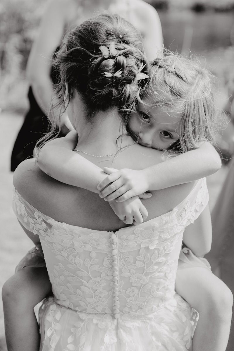 A woman in a lace wedding dress hugging a young girl in a sleeveless dress.