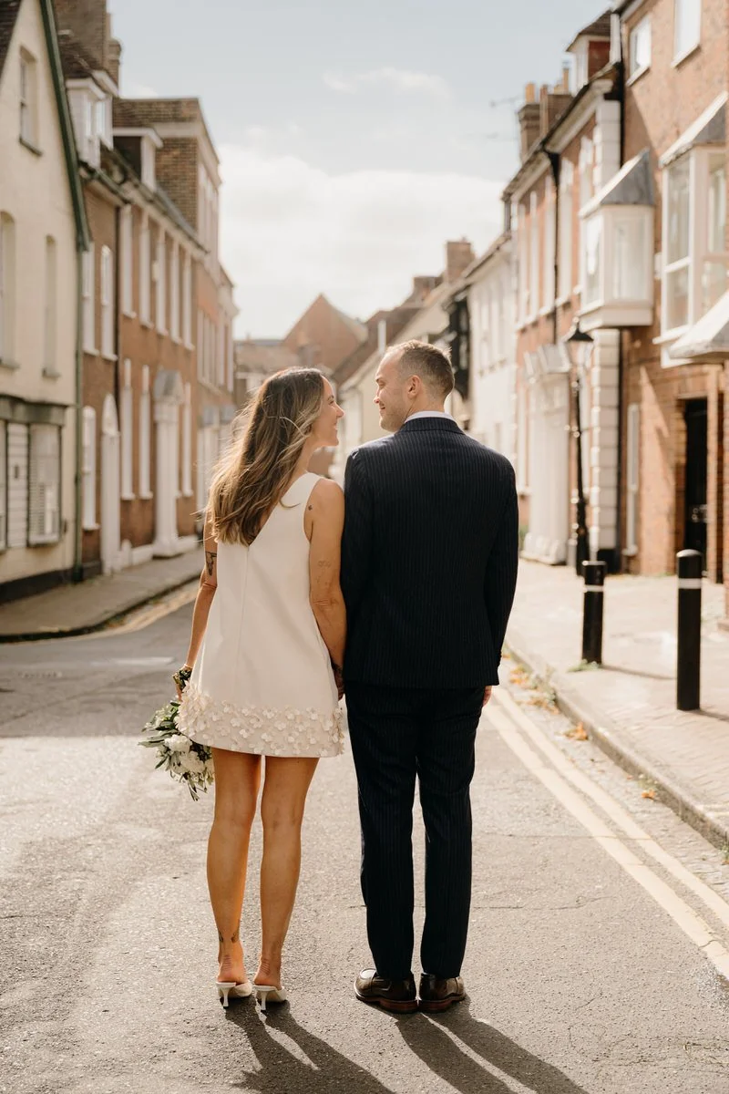 A newlywed couple holding hands and walking down a city street, smiling and looking at each other. The woman is in a white dress and heels, holding a bouquet, and the man is in a dark suit captured by Dorset Wedding Photographers pip&clo