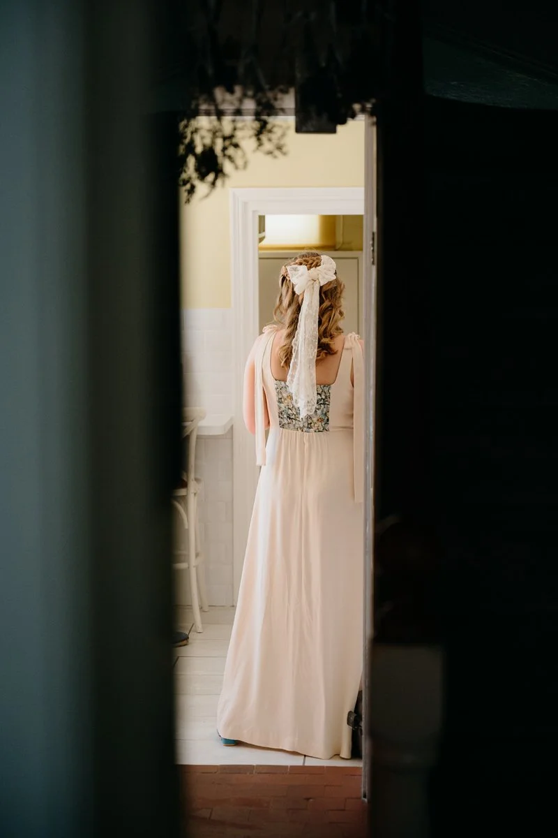 A bride in a long wedding dress with a lace back, standing in a doorway with a lace bow in her hair, seen from behind.
