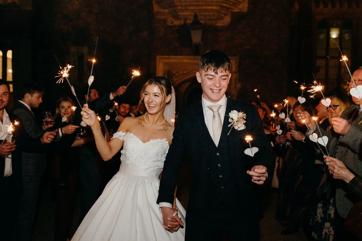 Bride and groom holding hands and smiling during wedding celebration with sparklers and guests in the background.