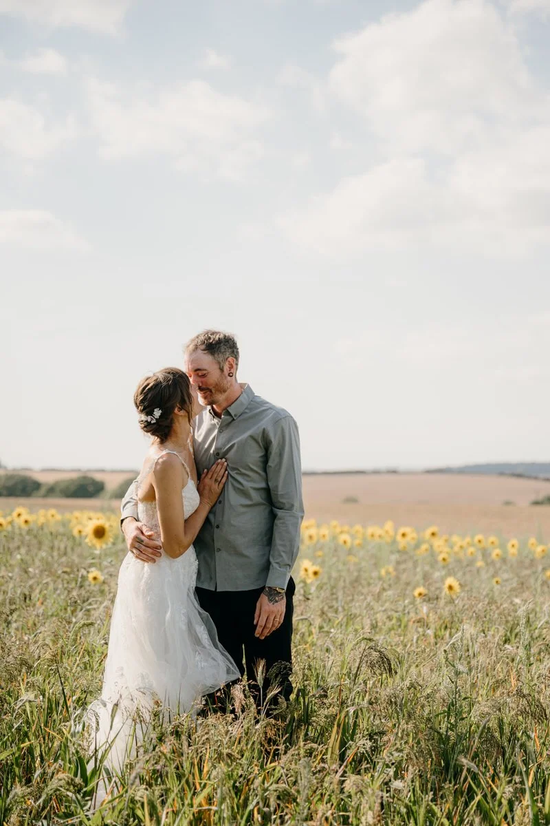 A newlywed couple, with the woman in a wedding dress and the man in a gray shirt, embracing in a sunflower field under a partly cloudy sky.
