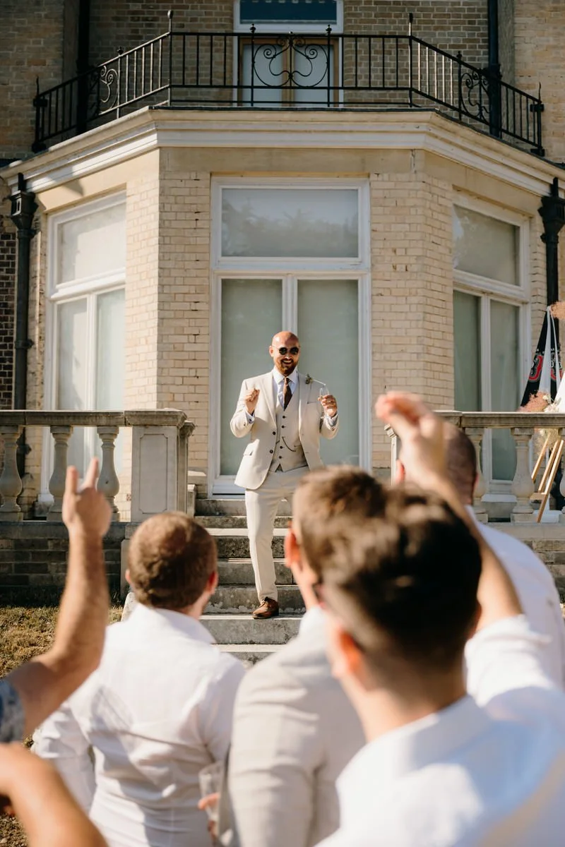 A man in a light-colored suit giving a speech to a group outdoors, with a building featuring large windows and a balcony in the background.