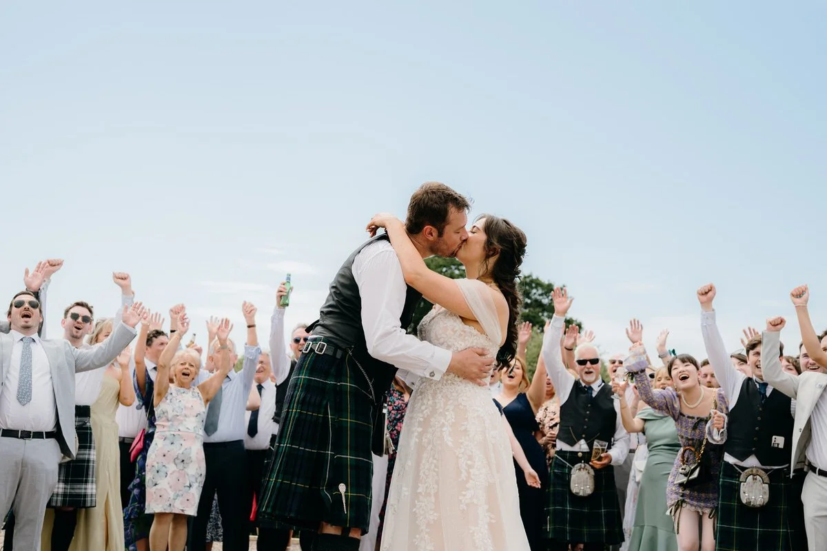 A newlywed couple kissing outdoors during their wedding celebration, surrounded by guests cheering and celebrating, with a clear sky in the background.
