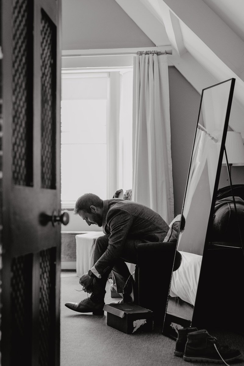 A man in a suit is sitting on a chair in a room, tying his shoelaces. The room has a window with curtains, a mirror leaning against the wall, and a bed with a blanket. The photo is in black and white. Captured by Dorset Wedding Photographers pip&clo