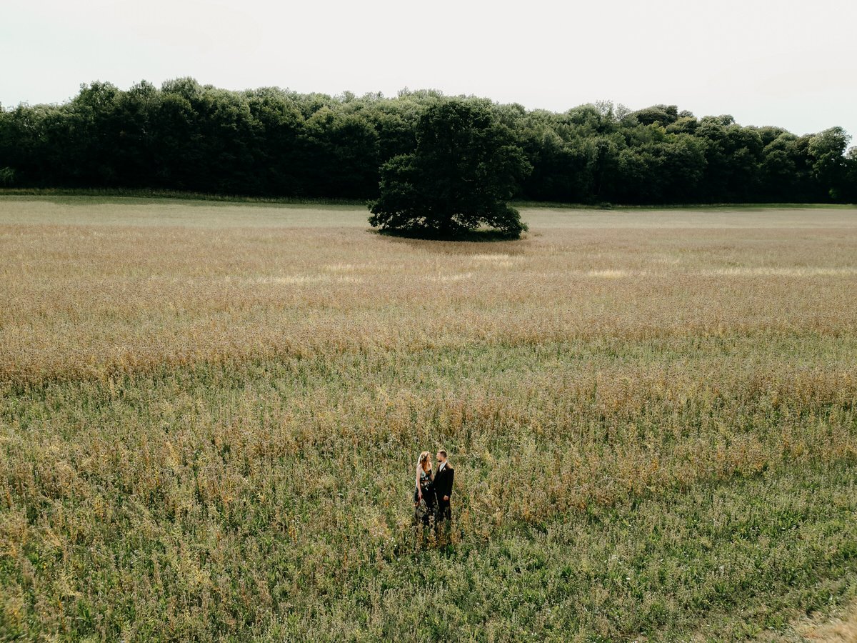 A couple standing in a grass field with a large tree and a forested area in the background.