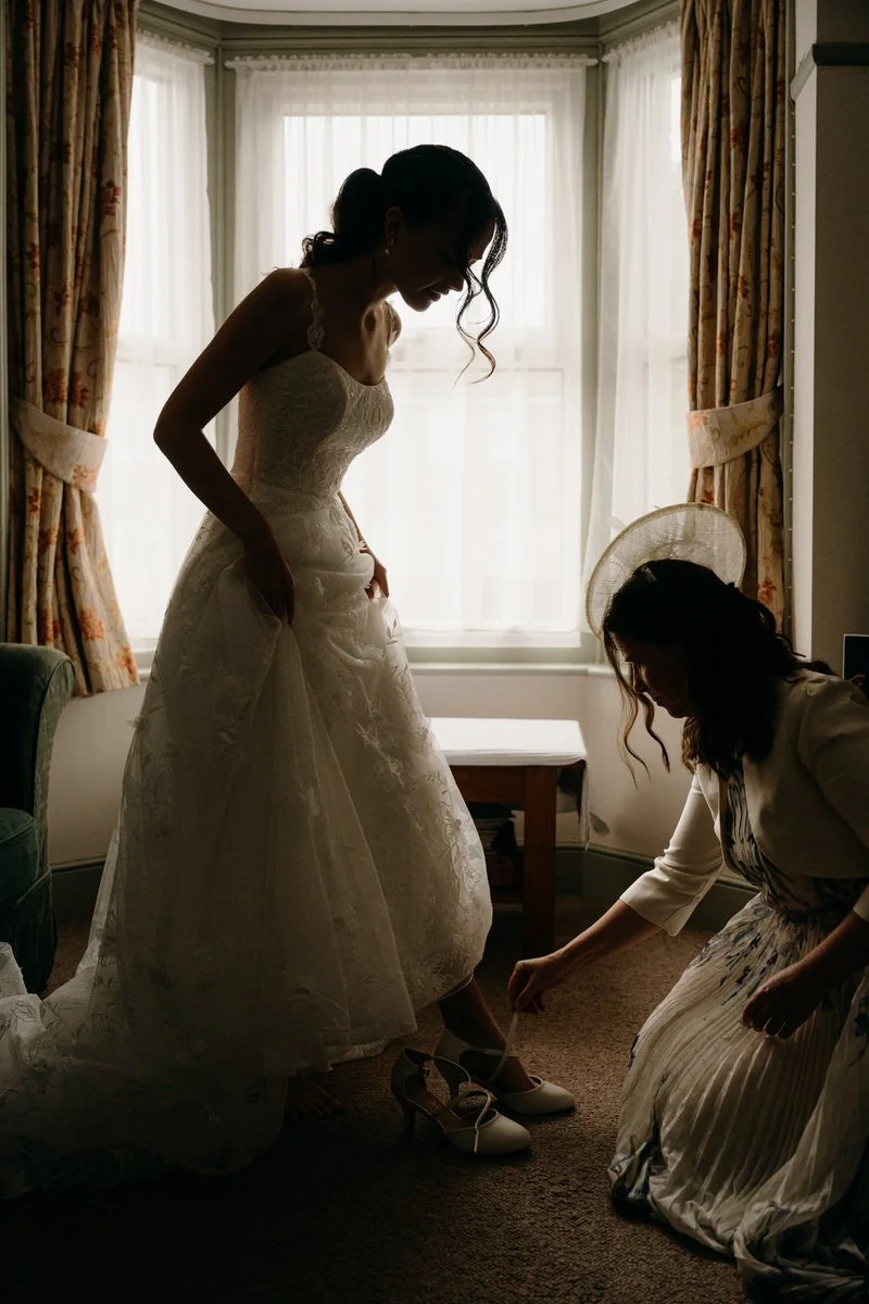 A bride in a wedding dress is helping herself put on shoes while another woman assists her inside a softly lit room with large windows and floral curtains.