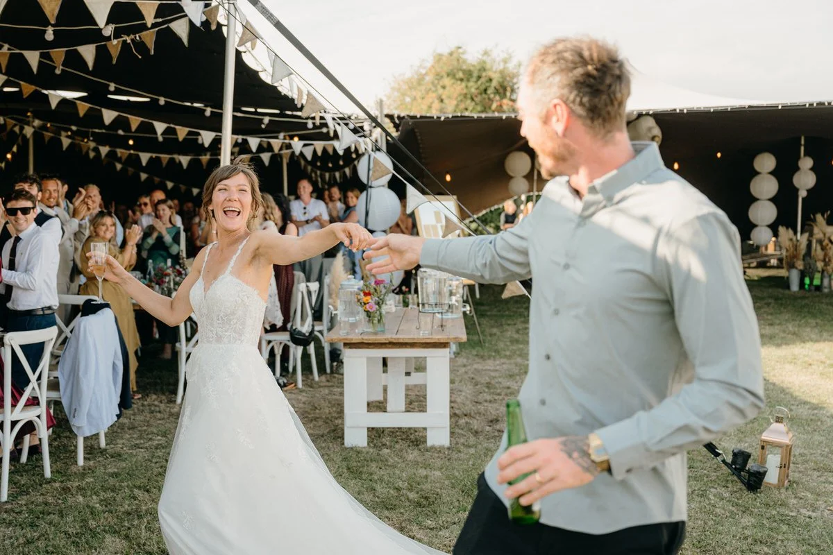 Bride and groom dancing at wedding reception outdoors, bride in white wedding dress holding a glass of champagne, groom in light shirt holding a drink, guests in the background clapping and smiling, decorated tent with bunting and paper lanterns.