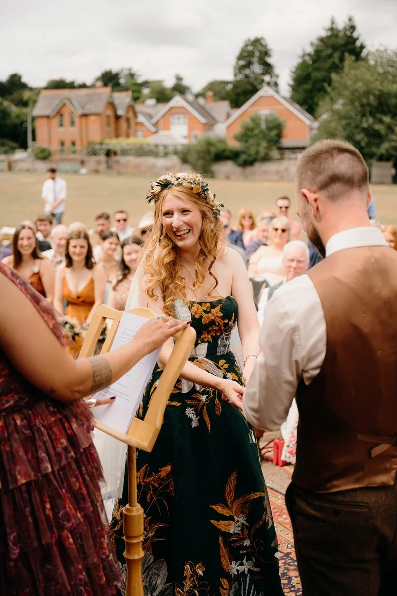 A bride in a green floral dress with long red hair and a flower crown smiling during her outdoor wedding ceremony photographed by Dorset wedding photographers pip&clo at a Dorset Village Hall Wedding.