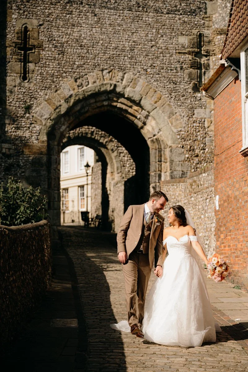 A bride and groom walking hand in hand on a cobblestone street with an ancient stone archway and brick buildings in the background during daytime. Captured by Dorset Wedding Photographers pip&clo