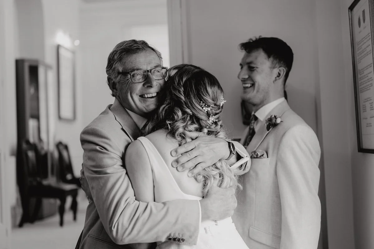 A heartfelt hug between three people, with a woman in a wedding dress embracing an older man and a young man in a suit, both smiling brightly, in an indoor setting.