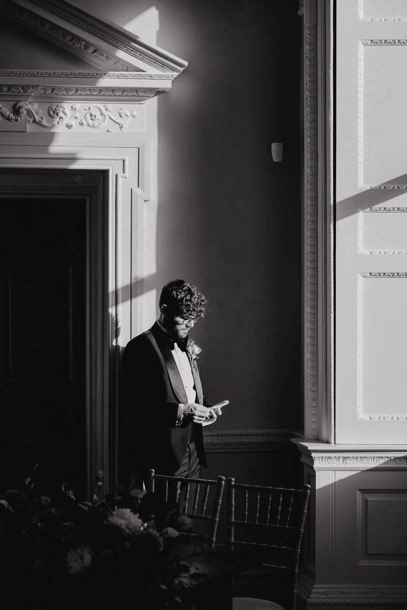 A man in a tuxedo with glasses, standing near a large window, looking at his phone. The room has ornate molding and intricate wall details, with sunlight casting shadows.