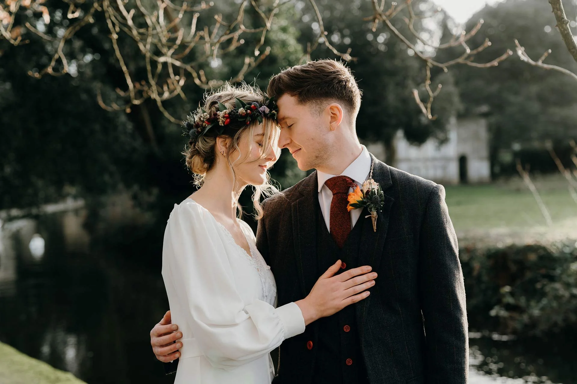 A bride and groom standing close with foreheads touching outdoors during daylight, with trees and a water body in the background, sharing a tender moment photographed by pip&clo Dorset wedding photographers in Christchurch Castle grounds.