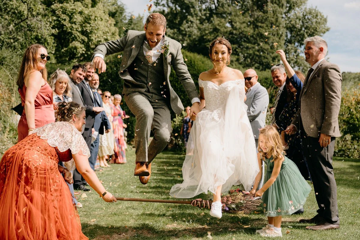 A bride and groom celebrating their wedding outdoors, jumping over a broom on a grassy field as wedding guests smile and watch.