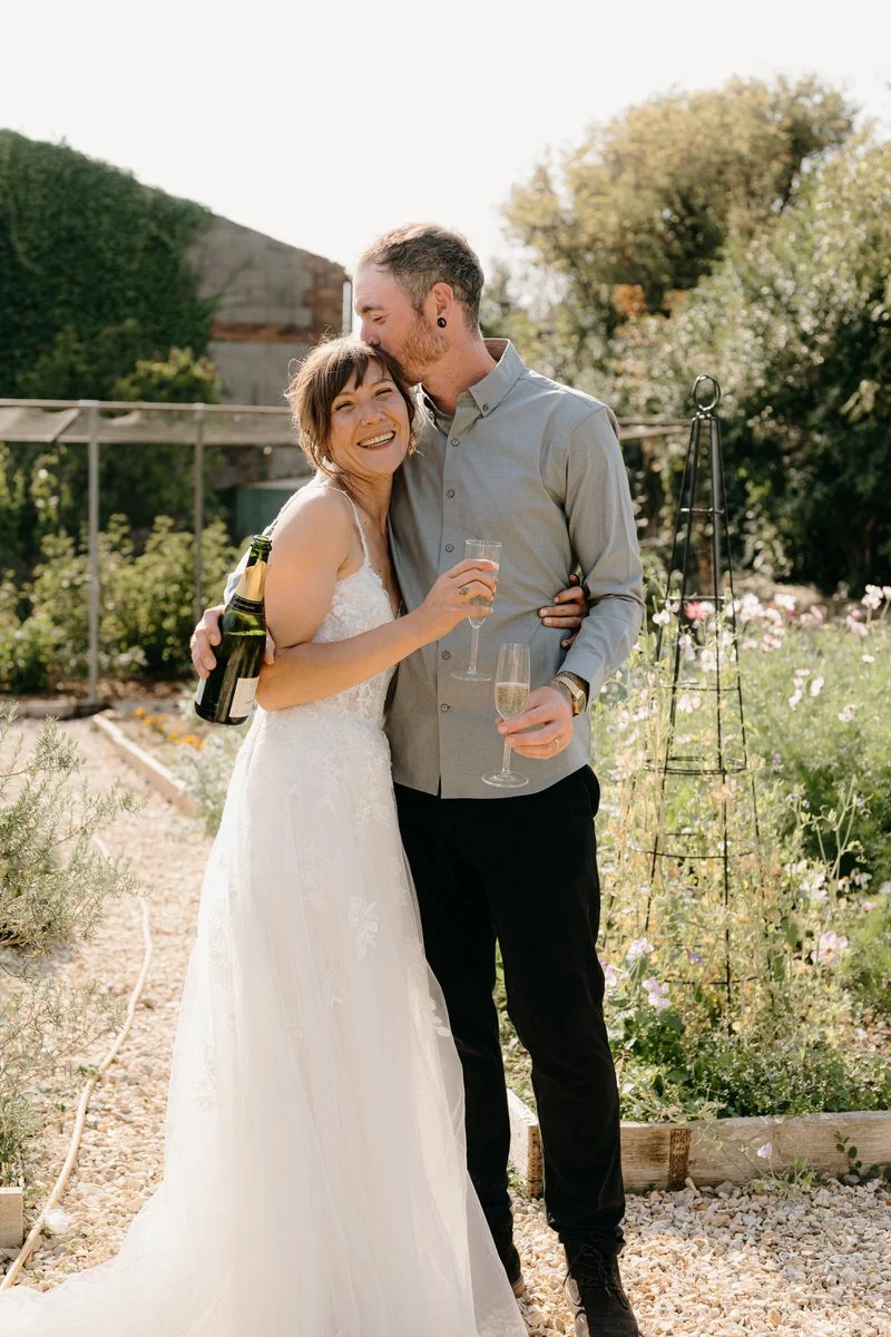 A married couple celebrating on a garden wedding day, with the woman in a white wedding dress holding a champagne glass, and the man in a gray shirt holding a champagne glass and a bottle photographed by Dorset wedding photographers pip&clo.