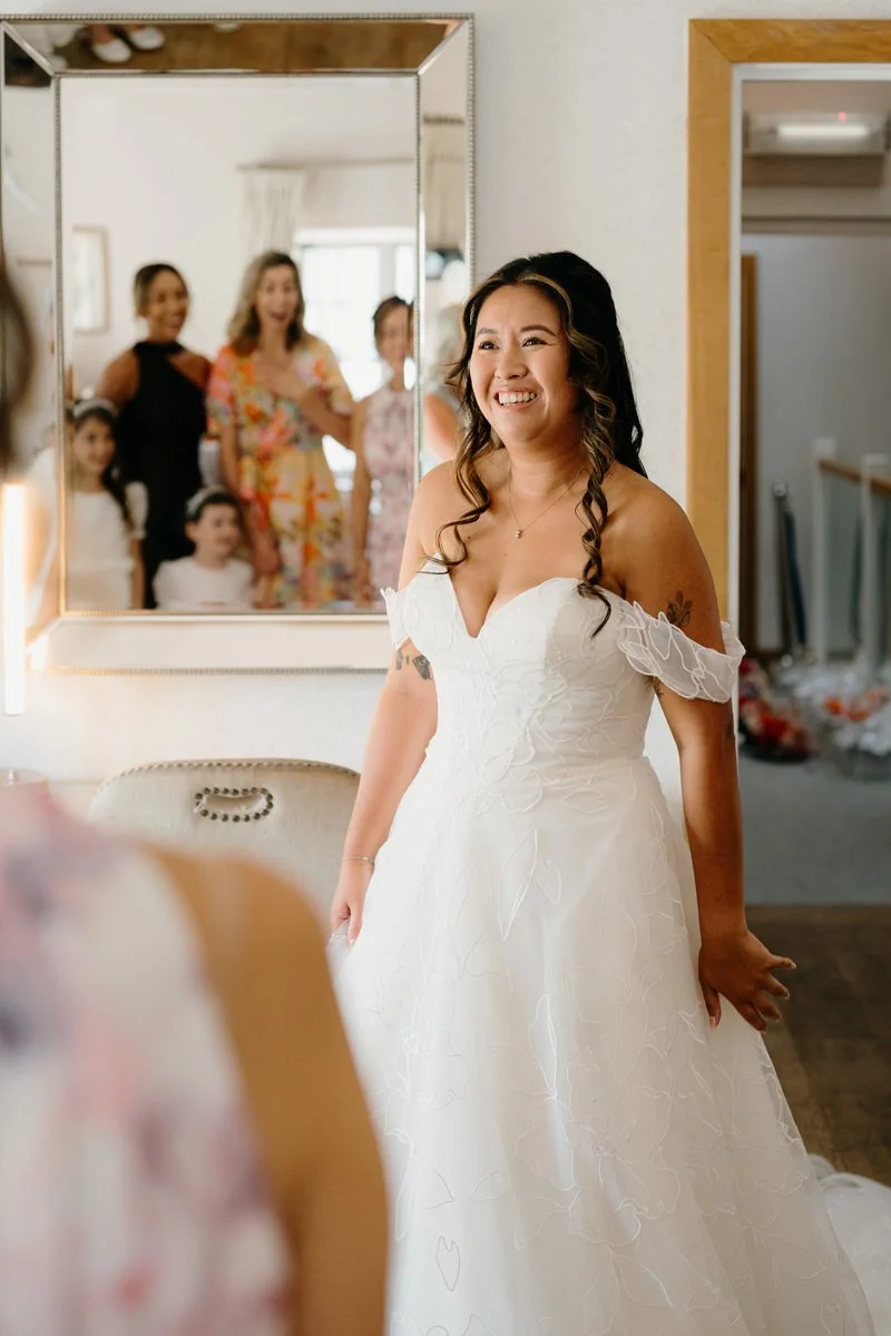 A bride wearing a white wedding dress with off-the-shoulder sleeves, smiling as she looks at her reflection in a mirror. In the background, a group of women and children are smiling and reacting to her appearance.