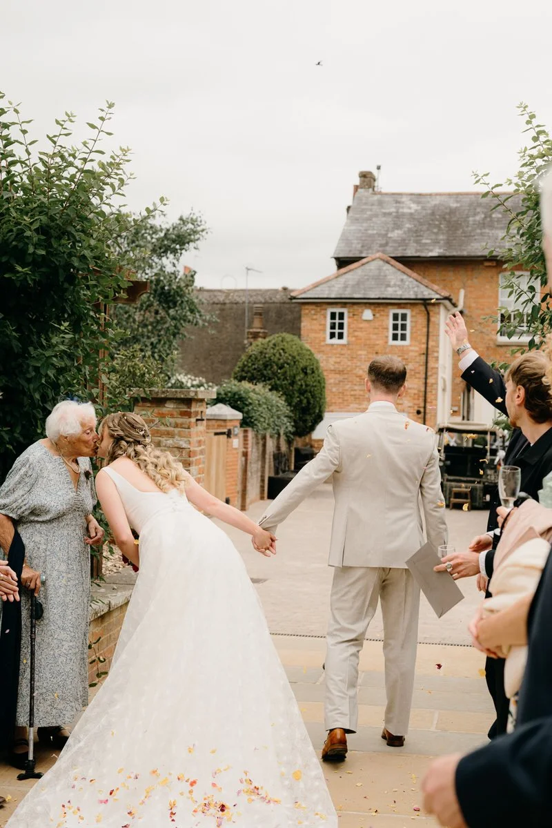 A wedding celebration with a bride and groom holding hands, greeting an elderly woman, while guests cheer and celebrate outside.