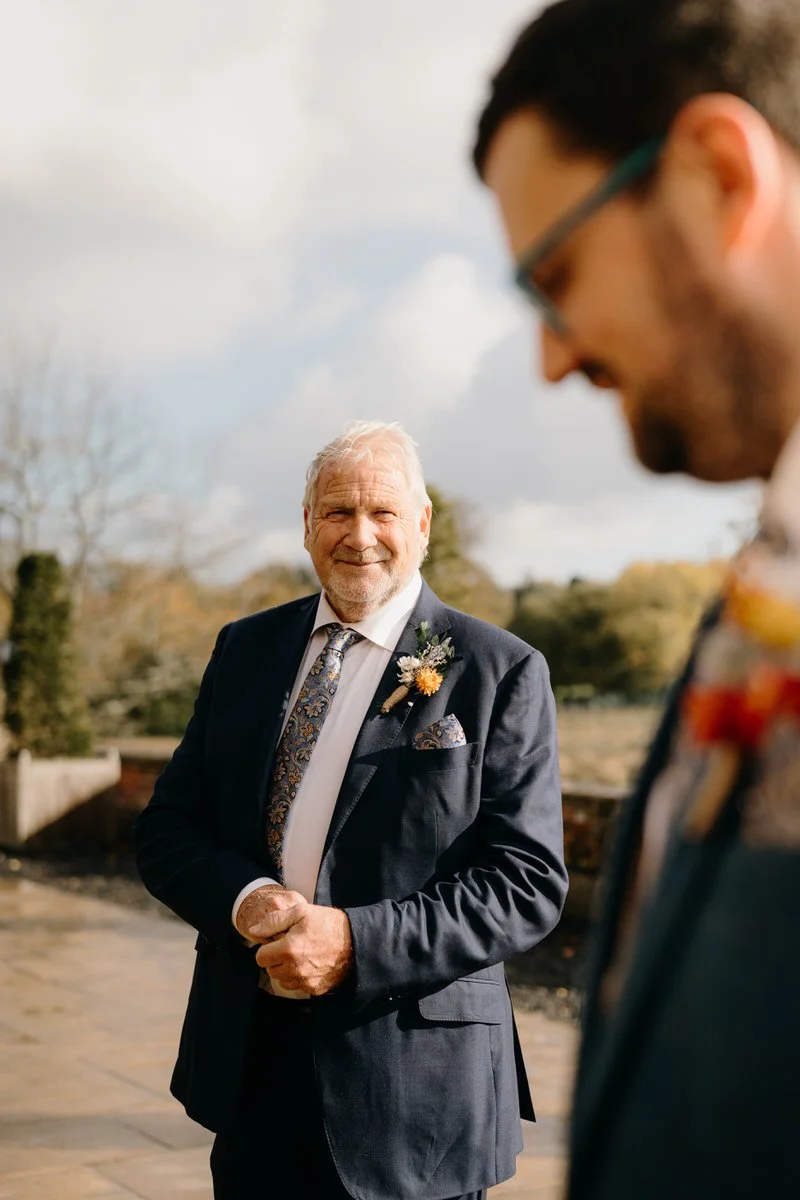 An elderly man in a suit with a boutonniere, smiling outdoors with a man in glasses blurred in the foreground, clouds, and trees in the background.