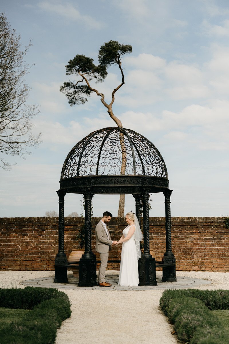 A bride and groom holding hands under a decorative black gazebo outdoors, with a tree behind them and a brick wall in the background.
