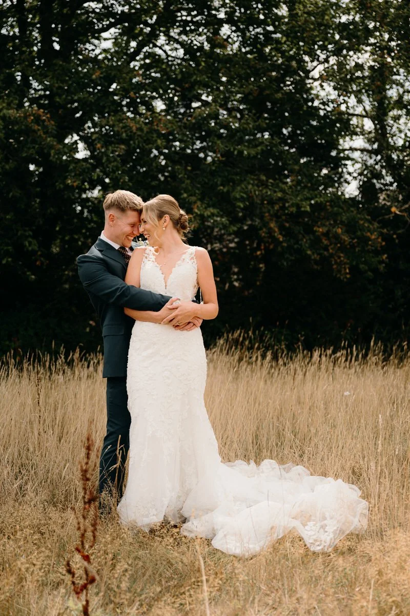 A newlywed couple standing close together in a field of tall grass with trees in the background, smiling and embracing each other.