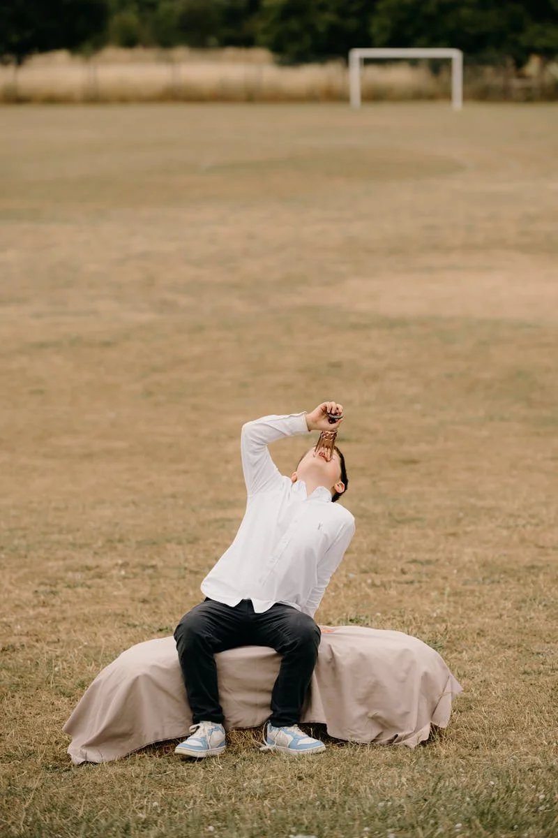 A person sitting on a beige-covered bench on a grassy field, drinking from a bottle, with a soccer goal in the background.