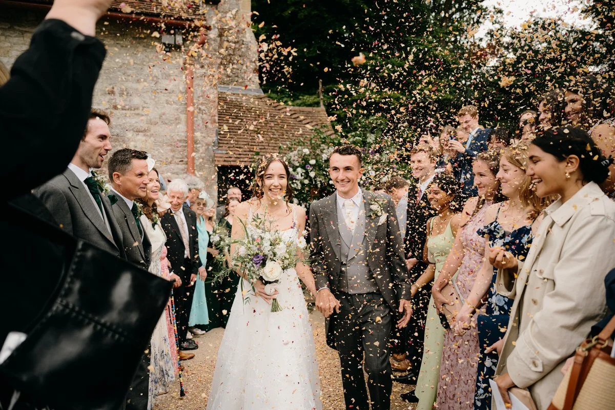 A bride and groom walking hand in hand through a crowd during a wedding celebration, with confetti being thrown and guests smiling.