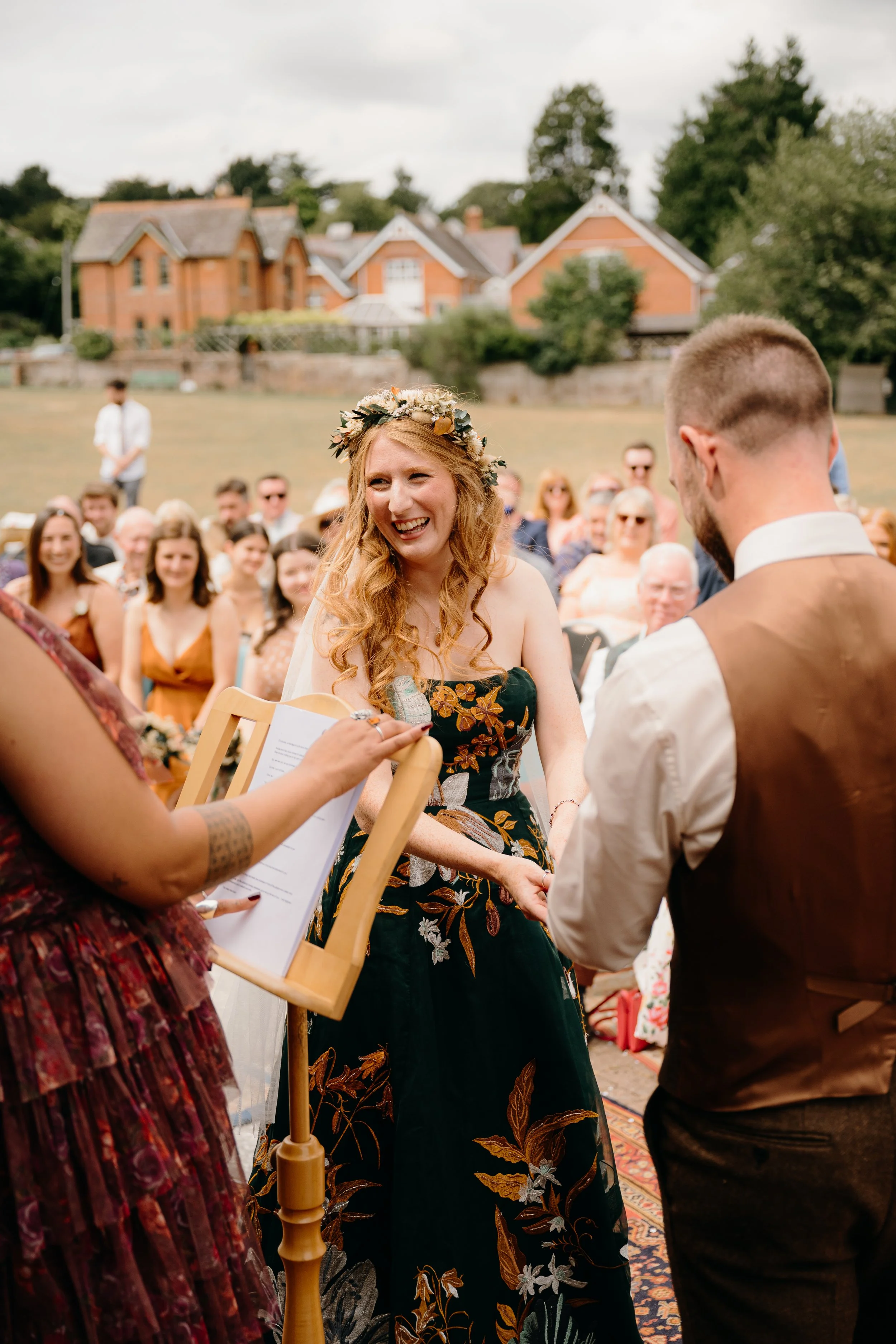 A bride with long, curly hair wearing a floral wedding dress and a flower crown smiling holding hands with her husband at an outdoor wedding ceremony. Guests seated in the background watch the moment. Captured by Dorset Wedding Photographers pip&clo