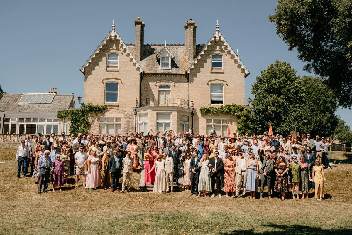 A large group of wedding guests dressed in formal and semi-formal attire posing outside a historic mansion in Dorset on a sunny day for a group photo, with a clear blue sky and green trees in the background captured by Dorset Wedding Photographers 
