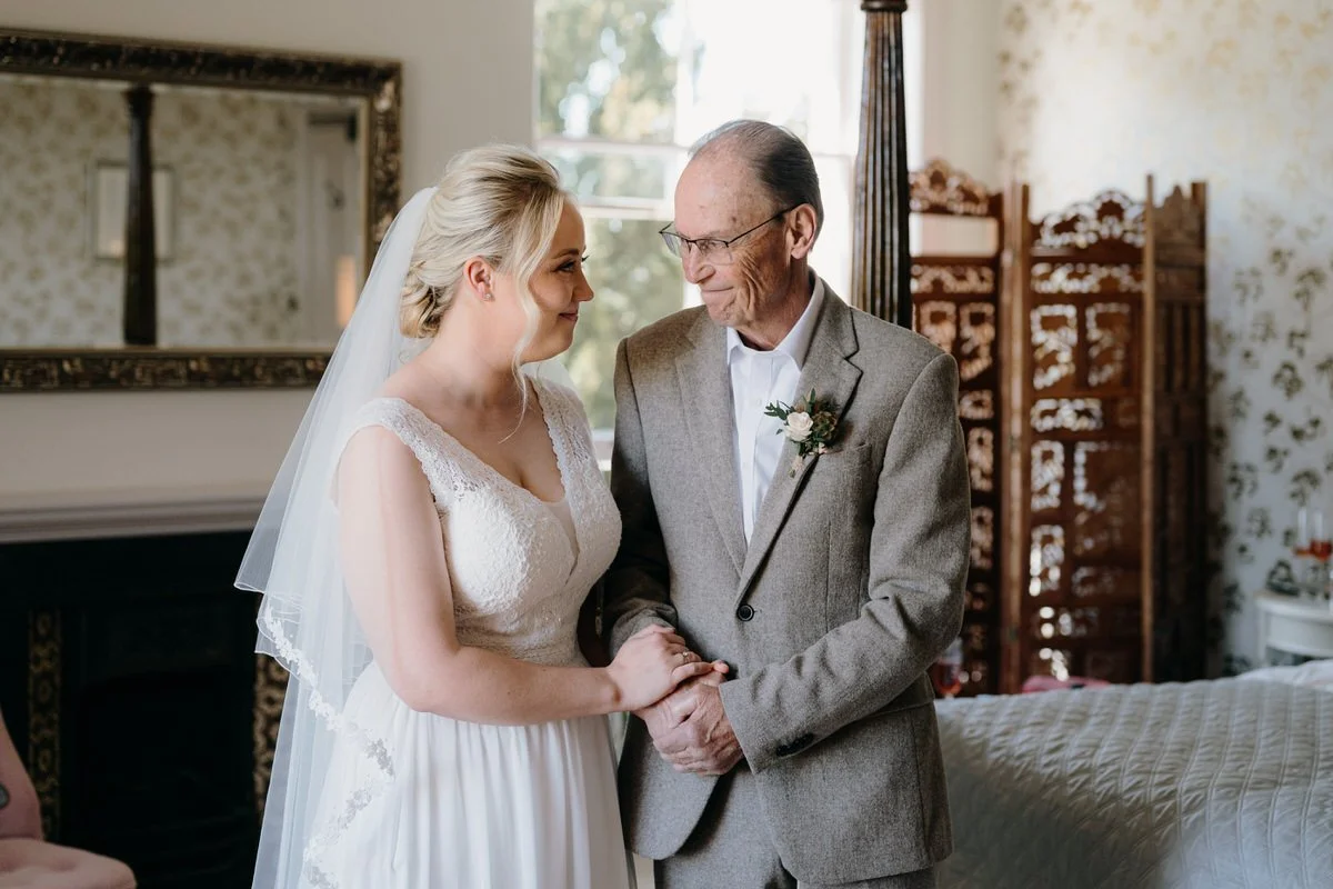 A bride and an older man, possibly her father, holding hands and smiling at each other in a warmly lit room during a wedding ceremony, with a mirror and decorative screen in the background.
