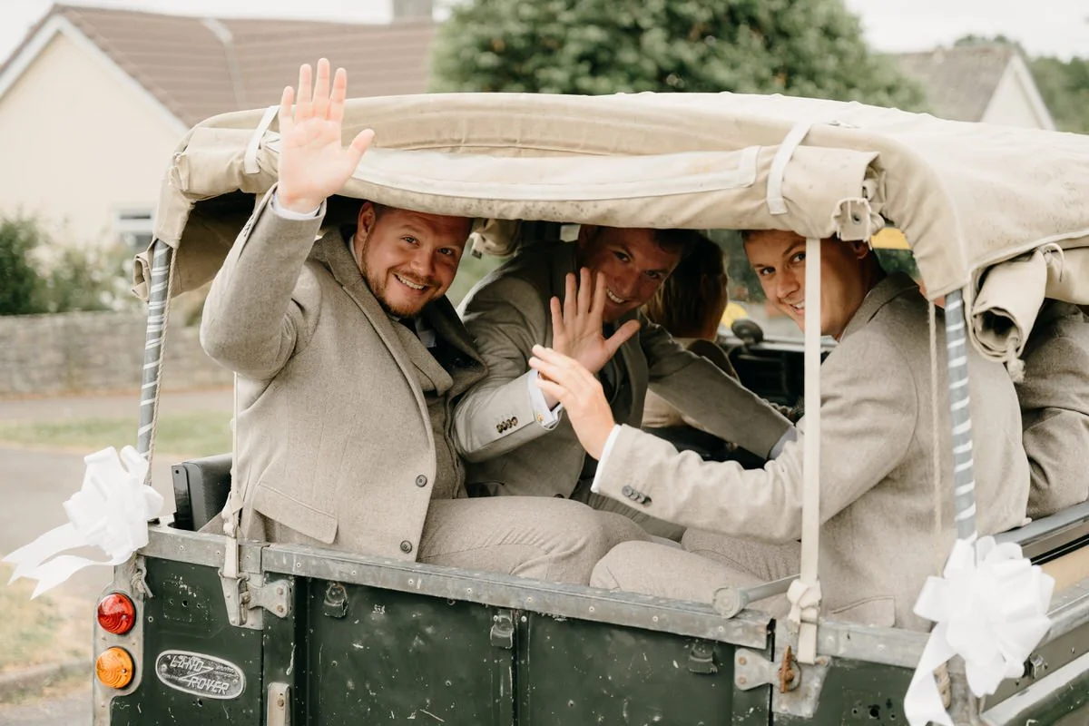 Grooms party in suits sitting in a decorated Landrover, smiling and waving at the camera.