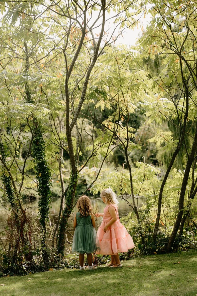 Two young girls in colorful dresses standing by a pond in a lush green park, surrounded by trees.
