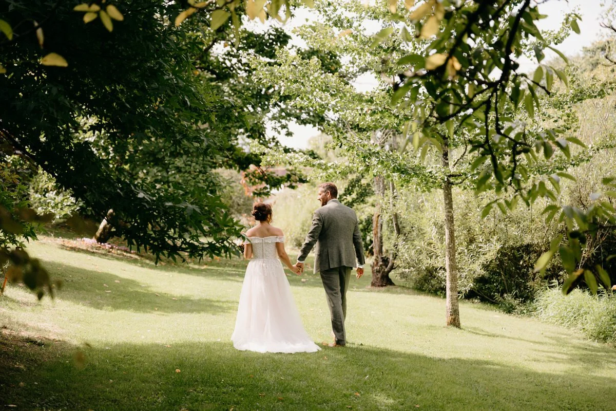 A bride and groom holding hands and walking through a lush green park or garden surrounded by trees and foliage on their wedding day.