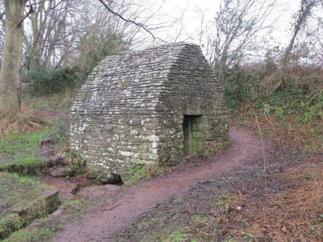 A small, ancient stone structure with an arched roof and a narrow doorway, situated in a wooded area with dirt paths and sparse vegetation.