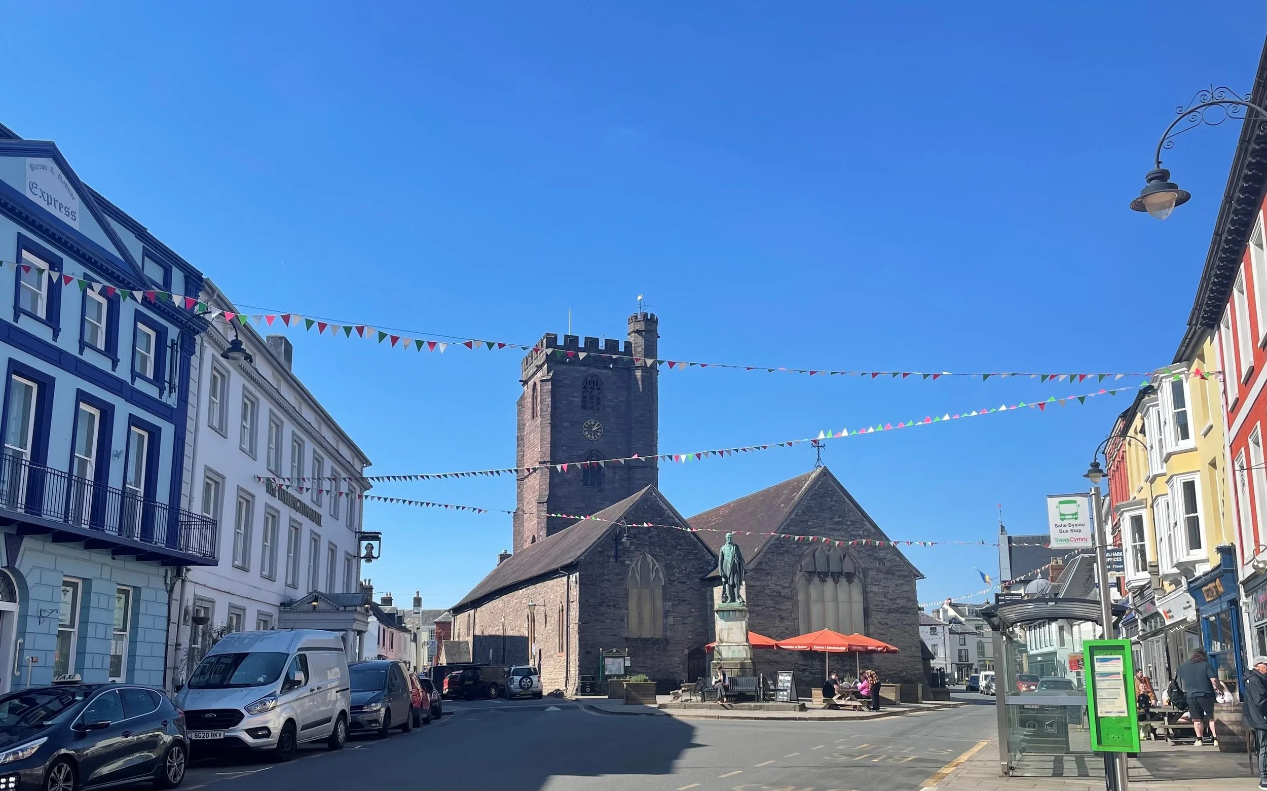 A historic church with a clock tower and statue in front, colorful triangular banners hang across a street with parked cars and pedestrians, in a sunny city setting.