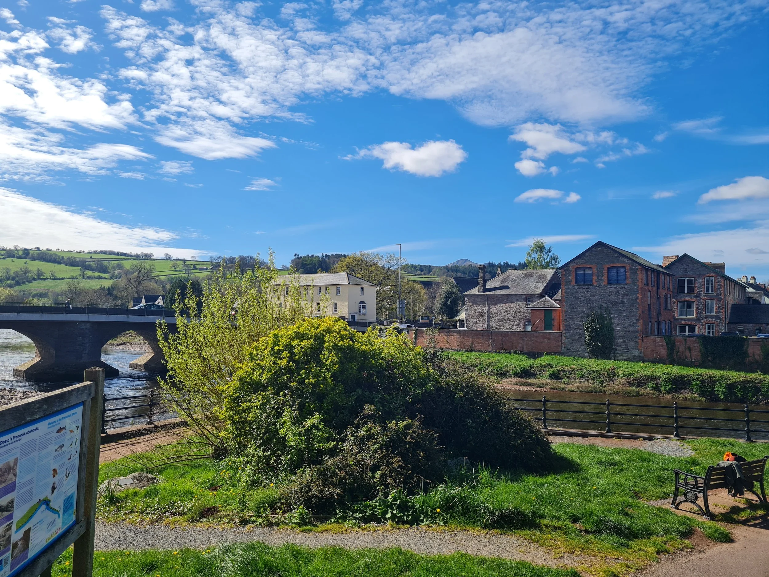 Scenic view of a small town by a river with a stone bridge, green hills, and partly cloudy blue sky.