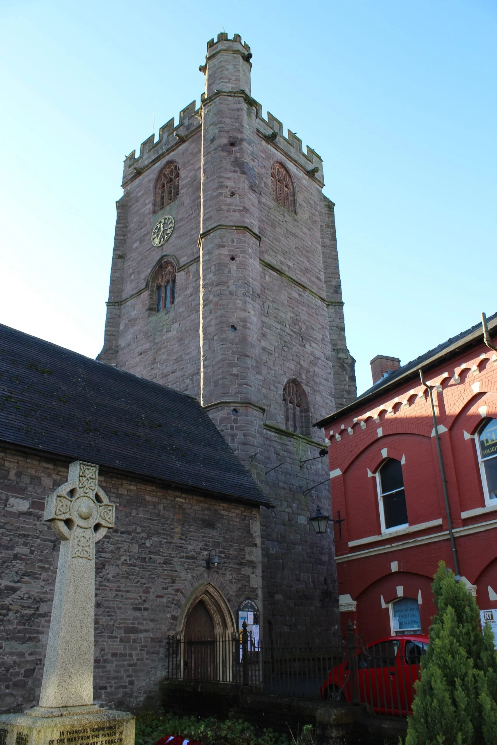 A tall stone church tower with a clock, flanked by a red brick building and a stone Celtic cross grave marker in the foreground.