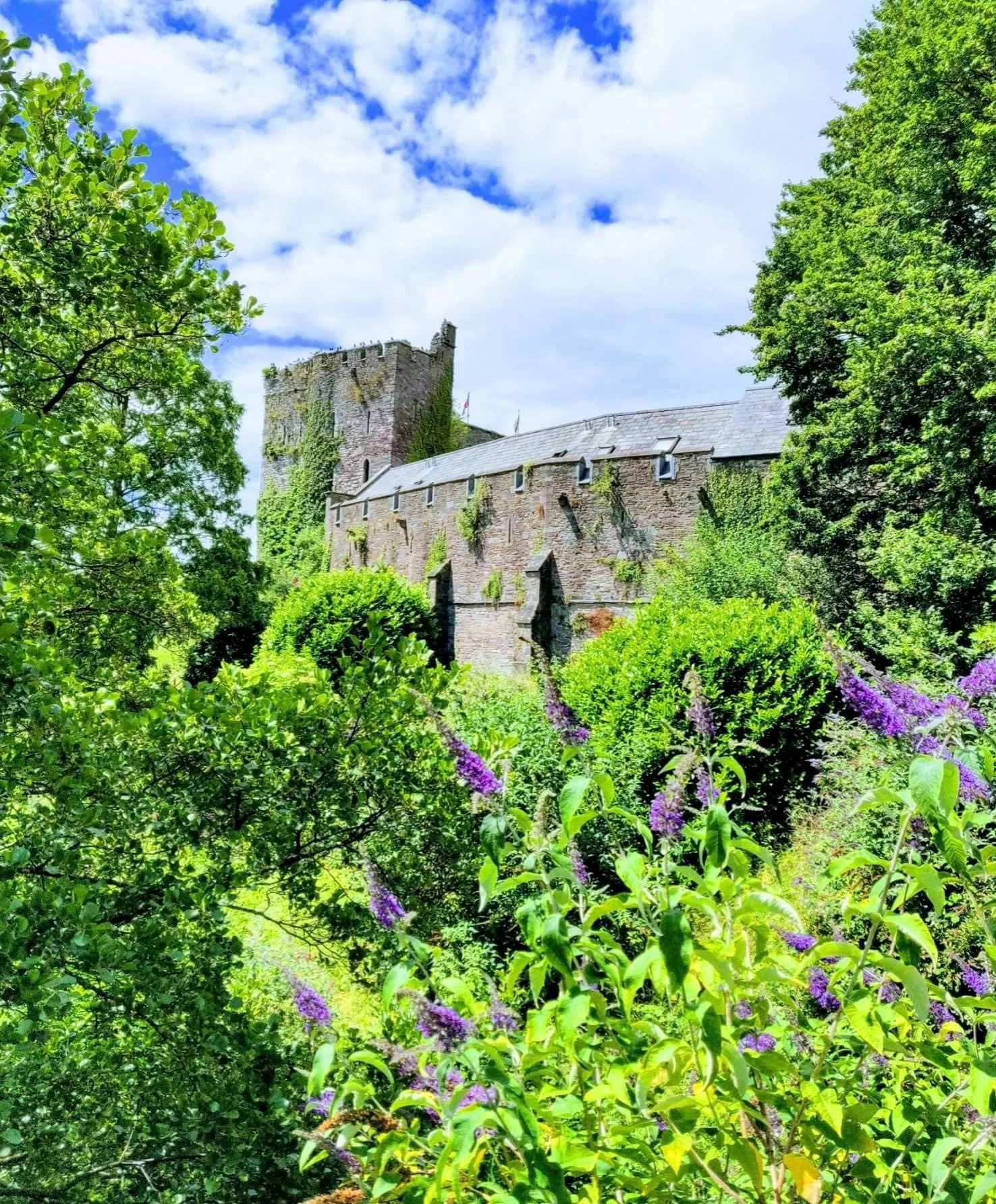 A stone castle surrounded by lush green trees and bushes with purple flowers beneath a partly cloudy sky.