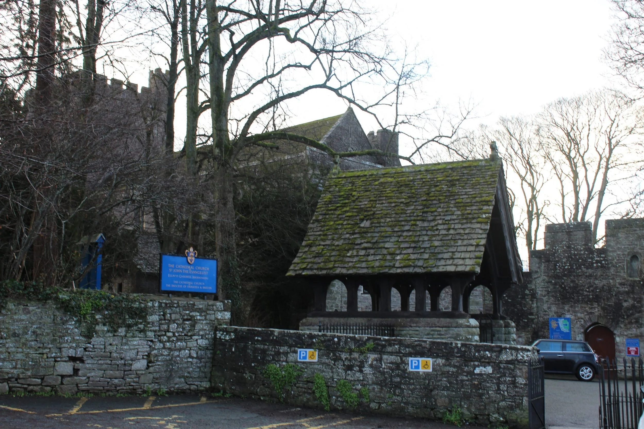 Historic stone church building with a moss-covered roof, surrounded by leafless trees, stone walls, and parking spaces in front.