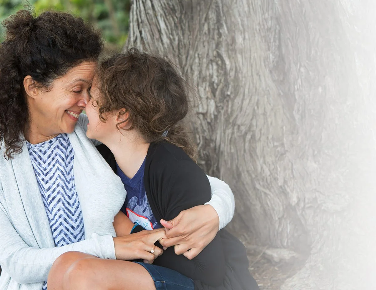 A mother and a young girl sharing a close moment outdoors, touching foreheads and smiling near a tree.