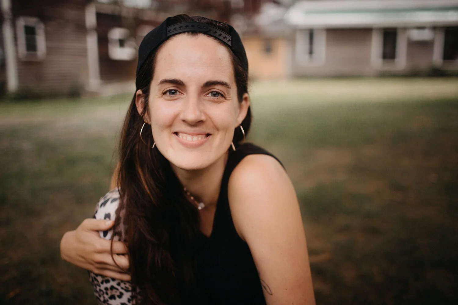 A young woman with long dark hair, wearing a black cap backwards, earrings, and a black sleeveless top, smiling outdoors with a blurred background of houses and grass.