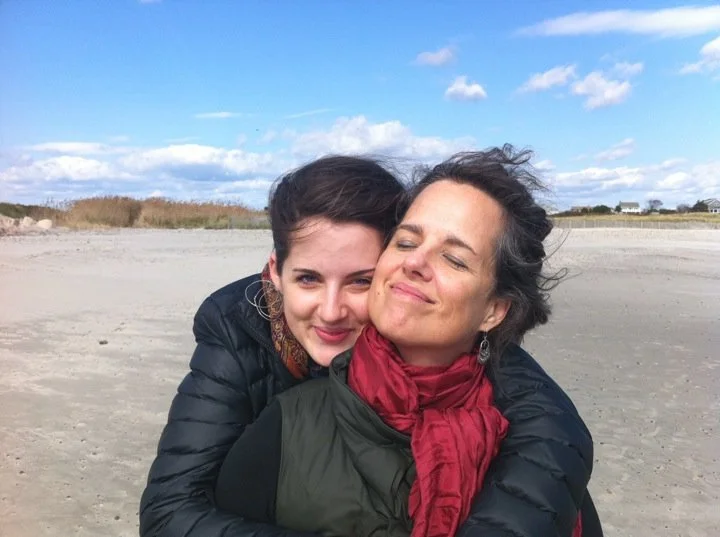 Two women hugging on a sandy beach with a cloudy blue sky and sparse vegetation in the background.